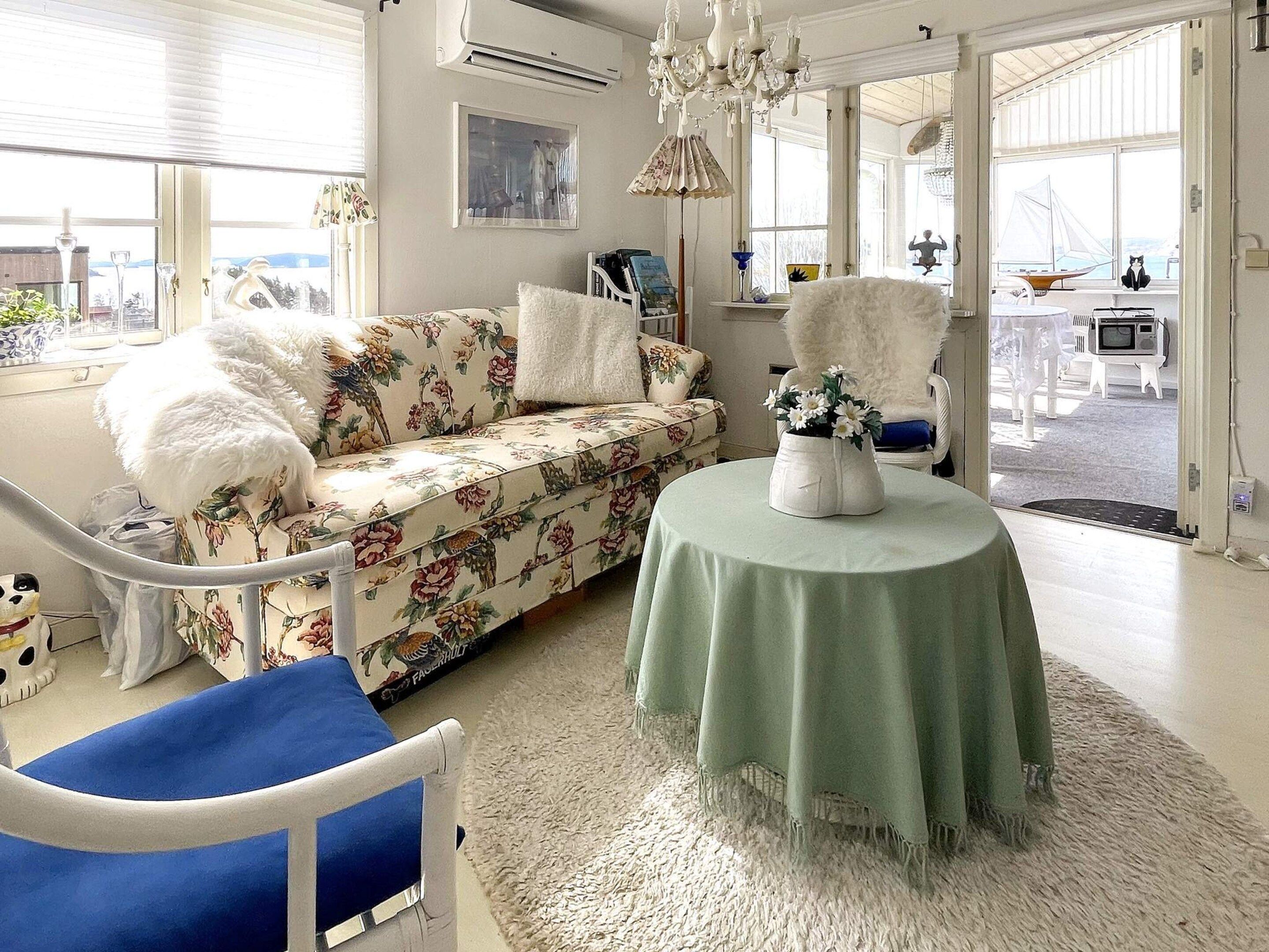 Een goed verlichte kamer met een bloemenbank, een witte, pluizige fauteuil, een ronde tafel met een vaas en uitzicht op buiten door grote ramen.