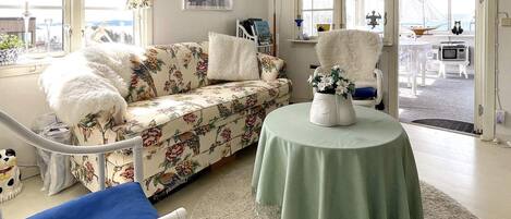 Una habitación luminosa con un sofá floral, un sillón blanco y mullido, una mesa redonda con un jarrón, y vistas al exterior a través de amplias ventanas.