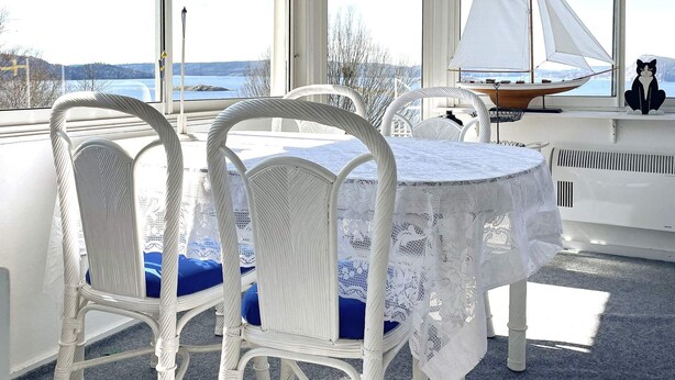 A room with a table and chairs, a sailboat model, and a view of a lake through large windows. - 4 Star Holiday Home in Uddevalla (Uddevalla)
