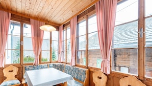 Une pièce en bois avec une grande table et des bancs, de grandes fenêtres ornées de rideaux roses, et une vue sur une maison à l’extérieur.