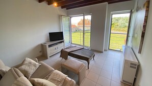 Ein Wohnzimmer mit Sofa, Couchtisch, Fernseher auf einem Holzboden und einer großen Schiebetür zum Balkon mit Blick auf Häuser und Grünflächen.
