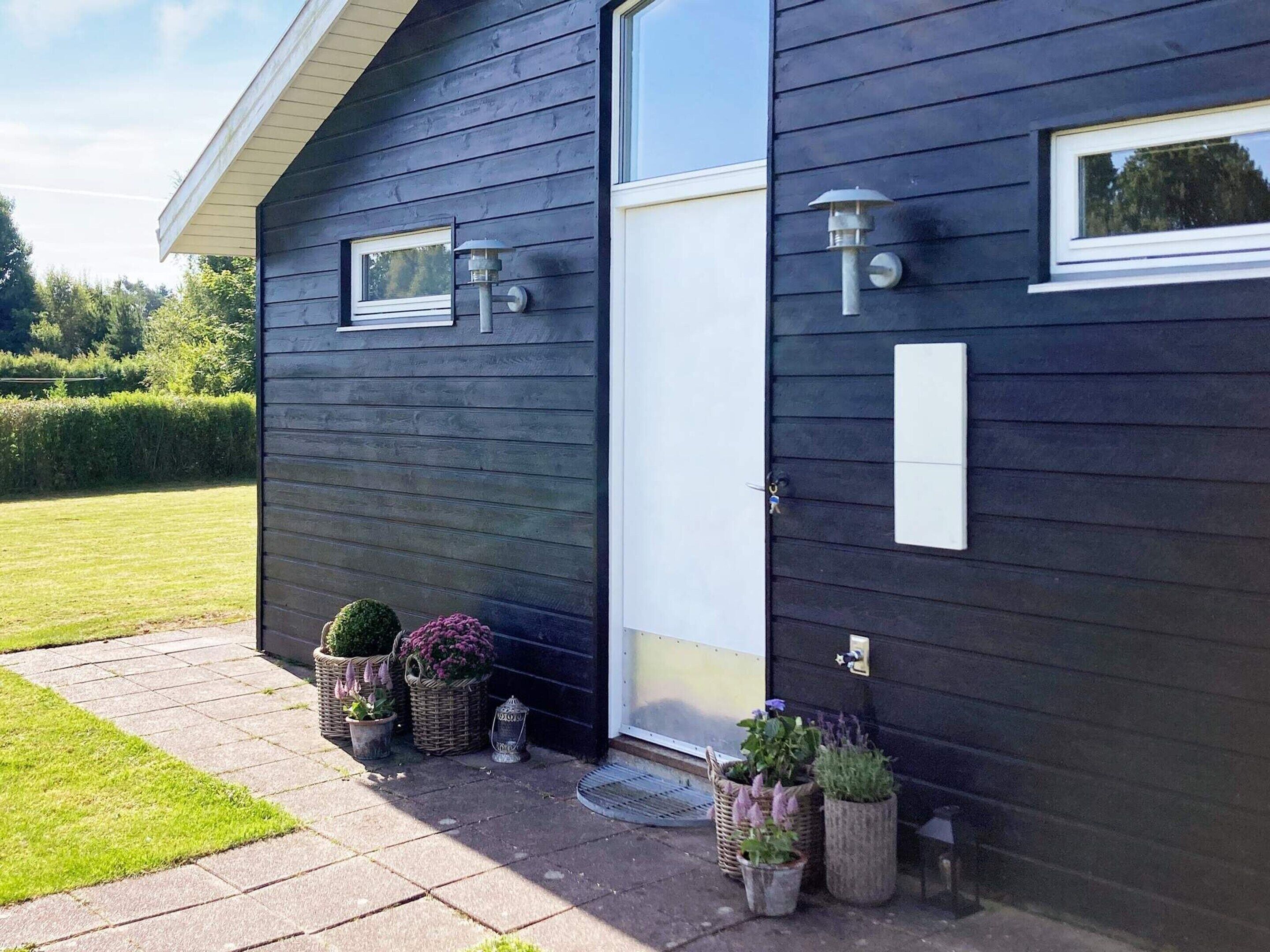A dark wooden building with a white door, two windows, and outdoor lighting fixtures. There are potted plants and a small garden area in front.