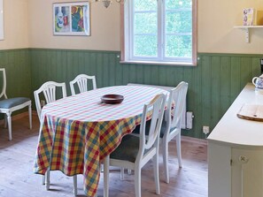 Un comedor con una mesa preparada para cuatro personas, rodeada de sillas blancas, un mantel de cuadros coloridos y una ventana con vistas a zonas verdes.