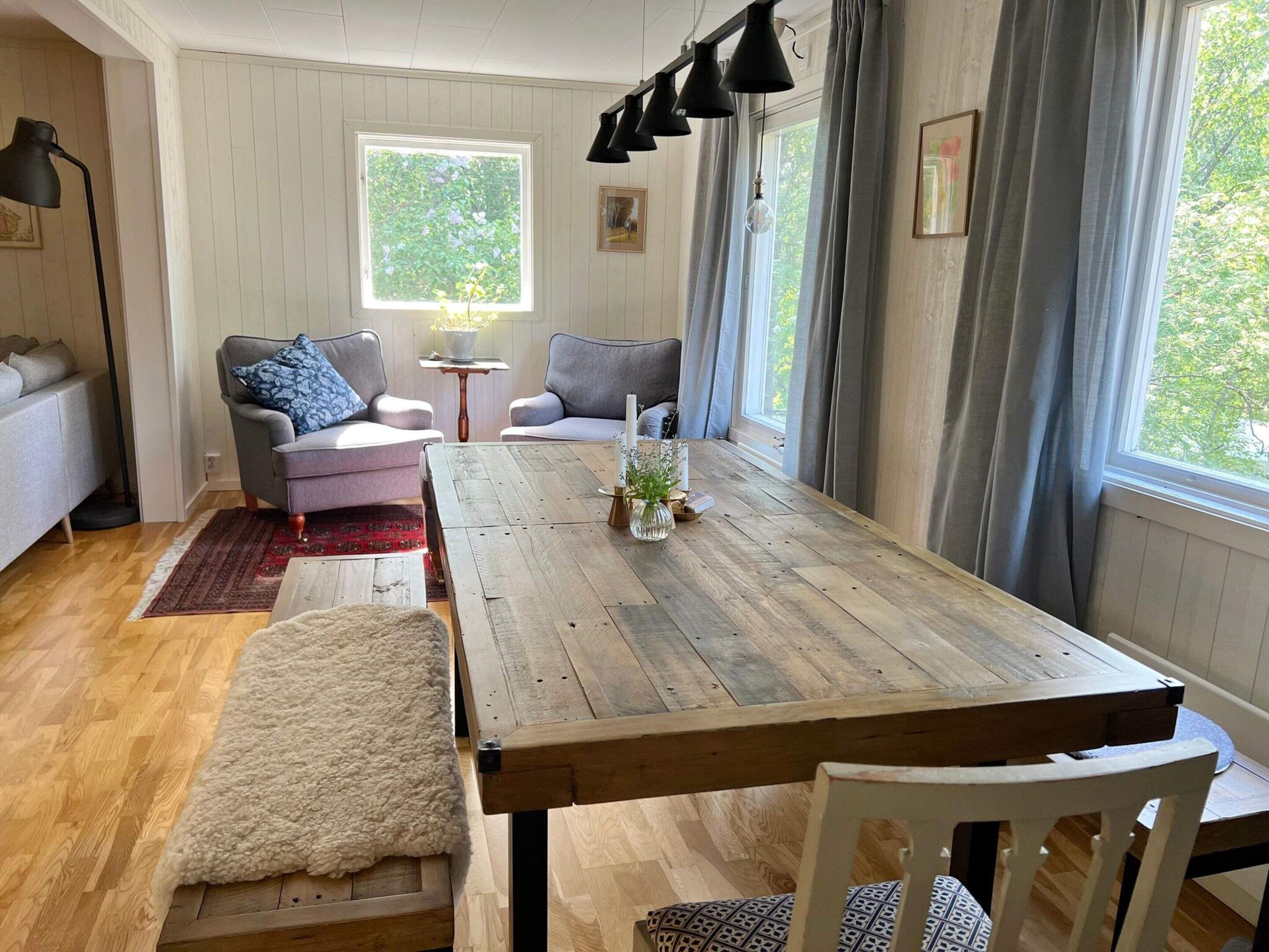 A cozy living room with a wooden dining table, two grey armchairs, a small side table, and a window with curtains.