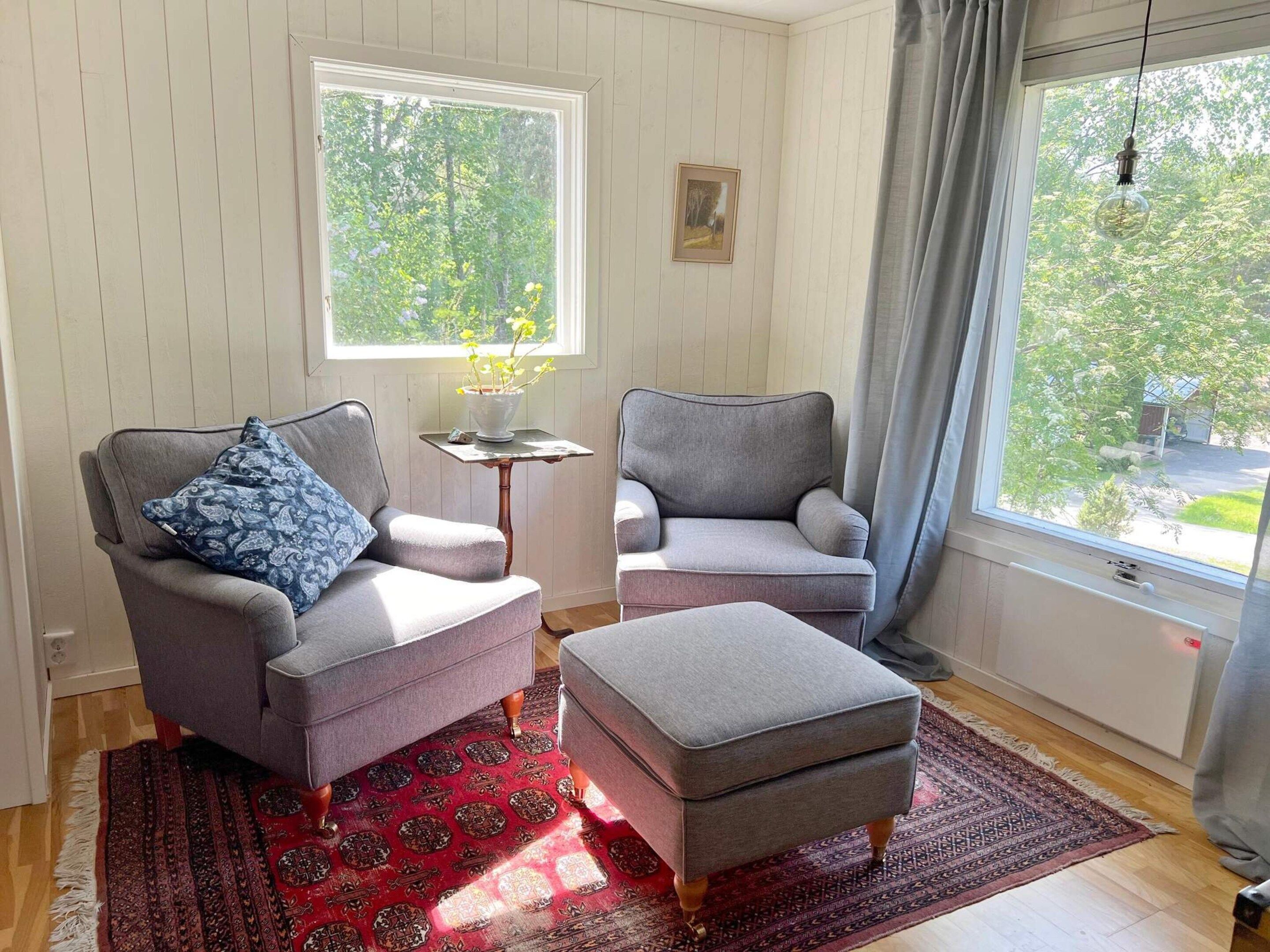 A room with two grey armchairs, a small table, a red patterned rug, and a window with a view of greenery.