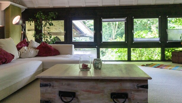 Una sala de estar con un sofá blanco, una mesa de centro de madera y ventanas grandes con vistas a zonas verdes.
