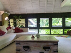 Un salon avec un canapé blanc, une table basse en bois et de grandes fenêtres donnant sur un paysage verdoyant.