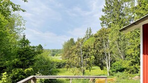A wooden deck with a metal railing overlooking a lush green landscape with trees and a clear sky. - 4 Person Holiday Home in Hallstavik (Hallstavik)