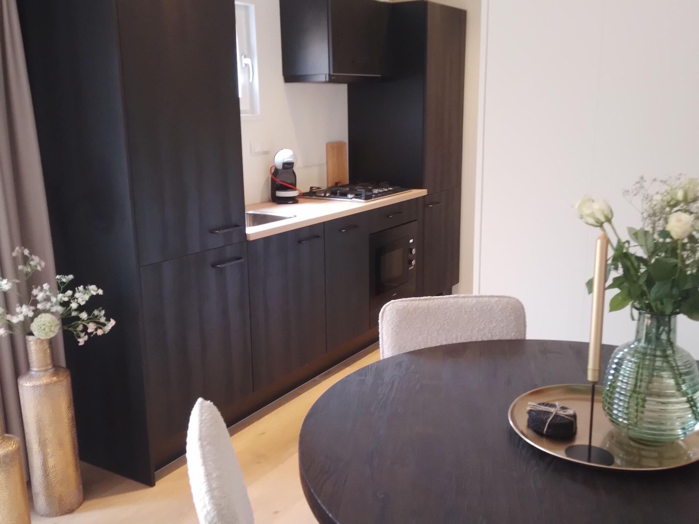 A modern kitchen with dark cabinetry, a white countertop, and a gas stove. There is a dining table with chairs and a vase with flowers.