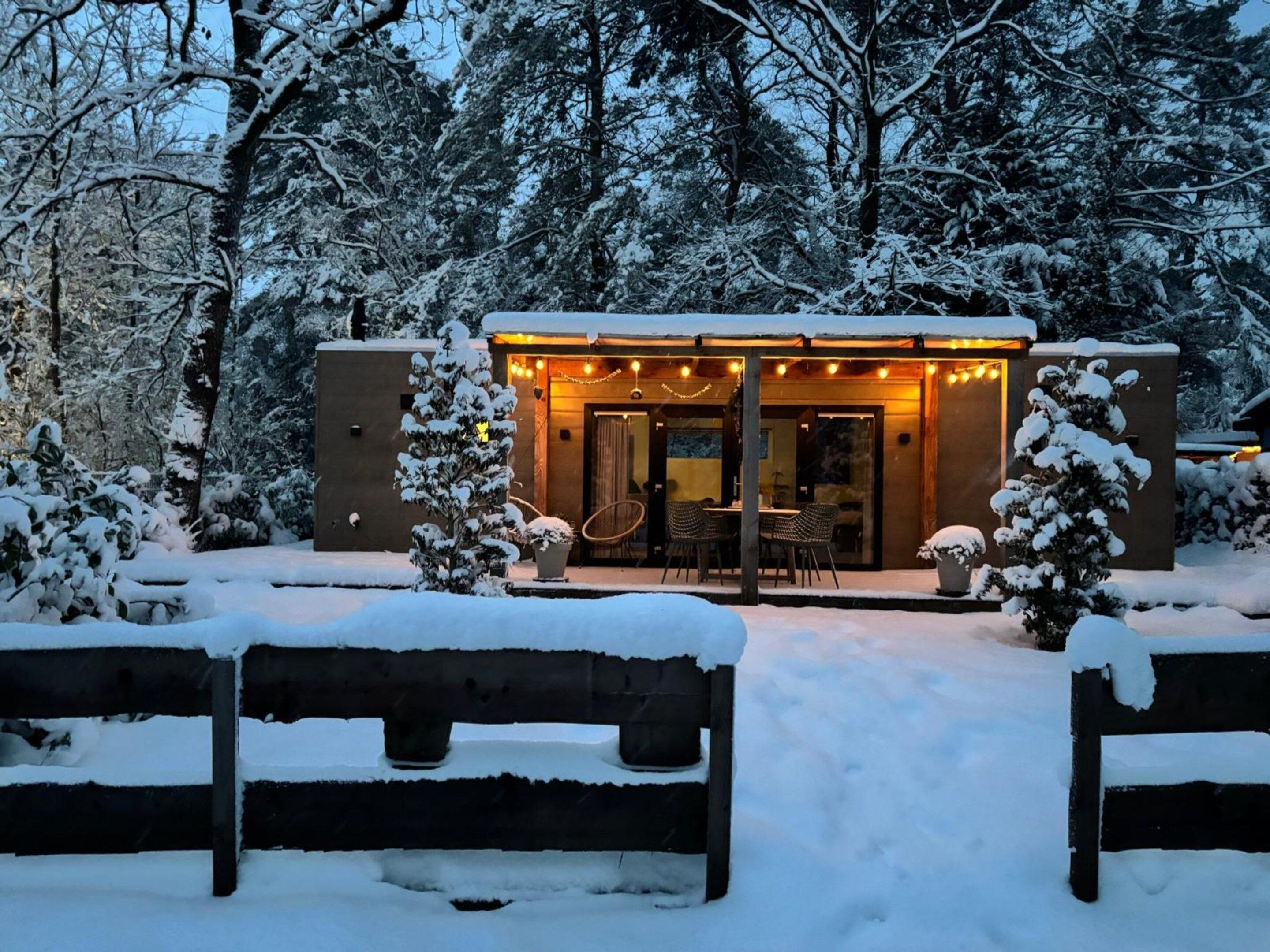 A cozy outdoor seating area with a table and chairs, surrounded by snow-covered trees and a building with warm lighting.