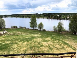Blick auf einen See mit einer Wiese, Bäumen und Häusern im Hintergrund.