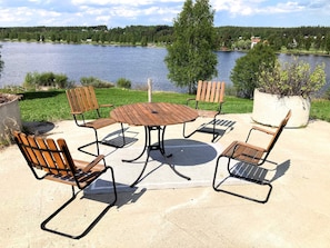 A patio with a round table and four wooden chairs, overlooking a lake with houses in the background. - 4 Person Holiday Home in Burtrask (Burträsk)
