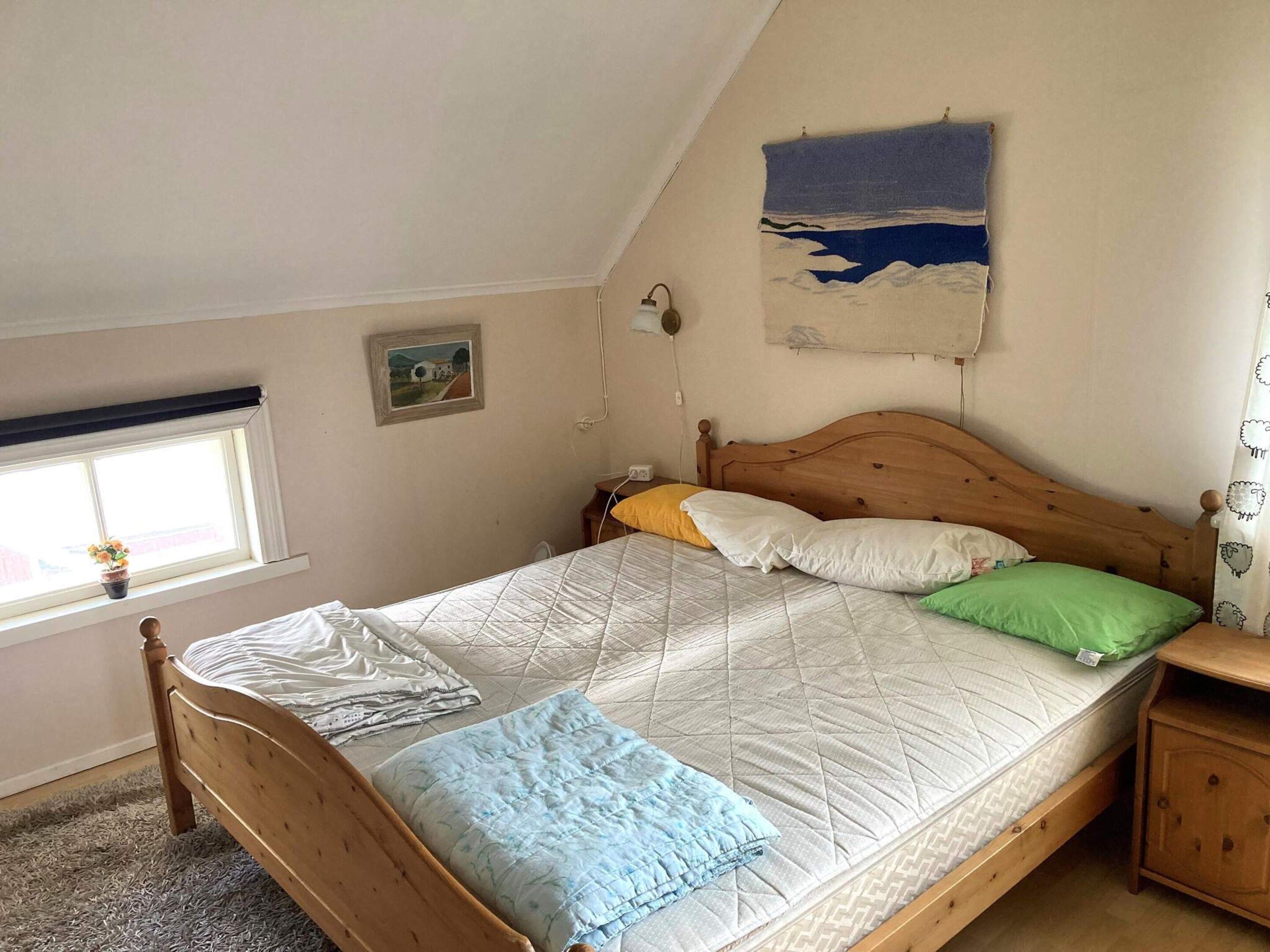 A single bed with a wooden frame, a headboard, and a quilted mattress. There is a bedside table with a lamp, a window with a view of the outside, and a wall-mounted painting.