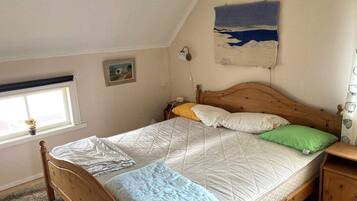 A single bed with a wooden frame, a headboard, and a quilted mattress. There is a bedside table with a lamp, a window with a view of the outside, and a wall-mounted painting.