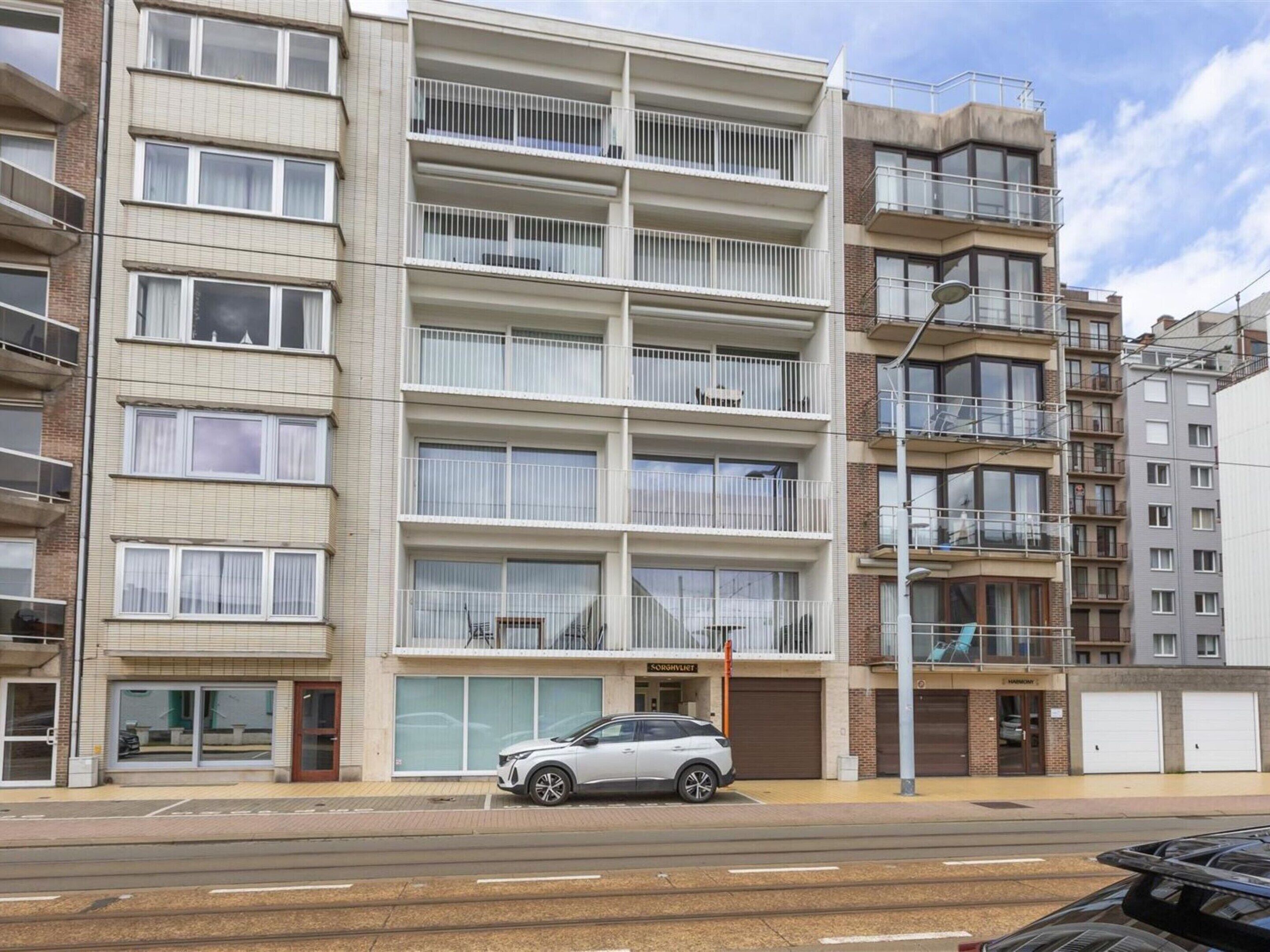 A multi-story residential building with balconies, a car parked in front, and a street view.