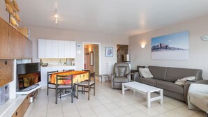 A modern living room with a sofa, coffee table, and a dining area with chairs and a table. There is a kitchen area with cabinets and a refrigerator. - Apartment in Middelkerke Near Sandy Beach (Middelkerke)