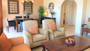 A living room with two beige armchairs, a wooden coffee table with fruit, a chandelier, and a dining area with a table and chairs. - Beach Penthouse Bermuda Beach 4 (Estepona)