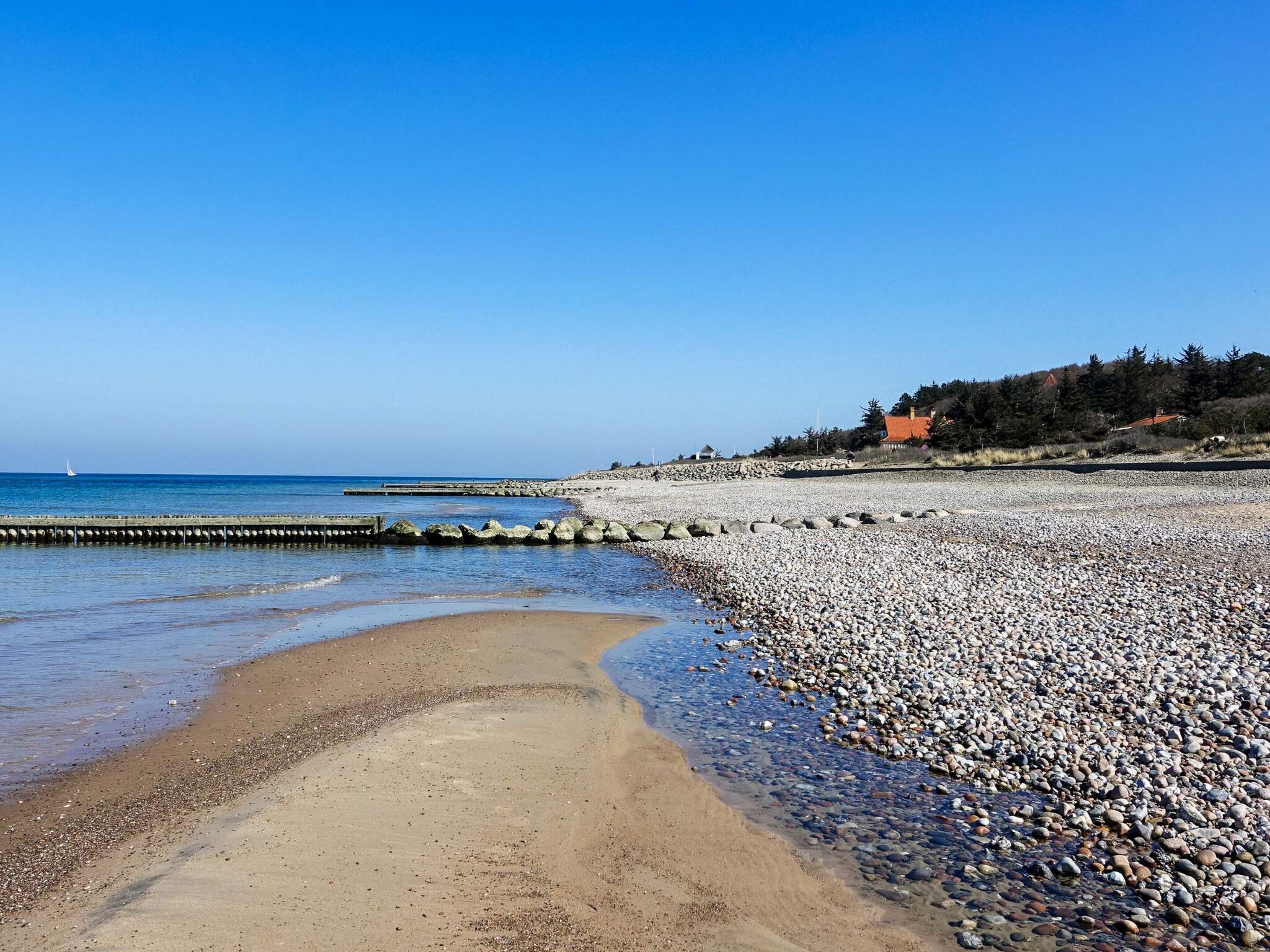 A pebbled beach with a clear sky, a distant sailboat, and a stone wall with a building in the background.