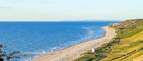 A coastal landscape with a pebble beach, grassy hills, and a clear view of the sea.