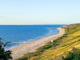 A coastal landscape with a pebble beach, grassy hills, and a clear view of the sea.