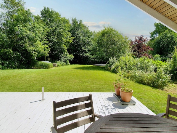 A wooden deck with a table and chairs, surrounded by a lush green lawn and various plants. - 6 Person Holiday Home in Graested (Graested)