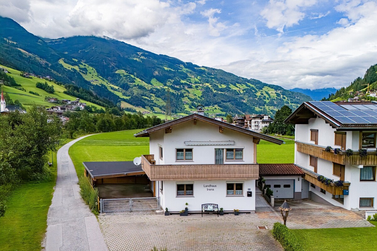 Ein zweistöckiges Gebäude mit der Aufschrift 'Landhaus Irene' an der Vorderseite, einer gepflasterten Auffahrt und Blick auf grüne Felder und Berge im Hintergrund.