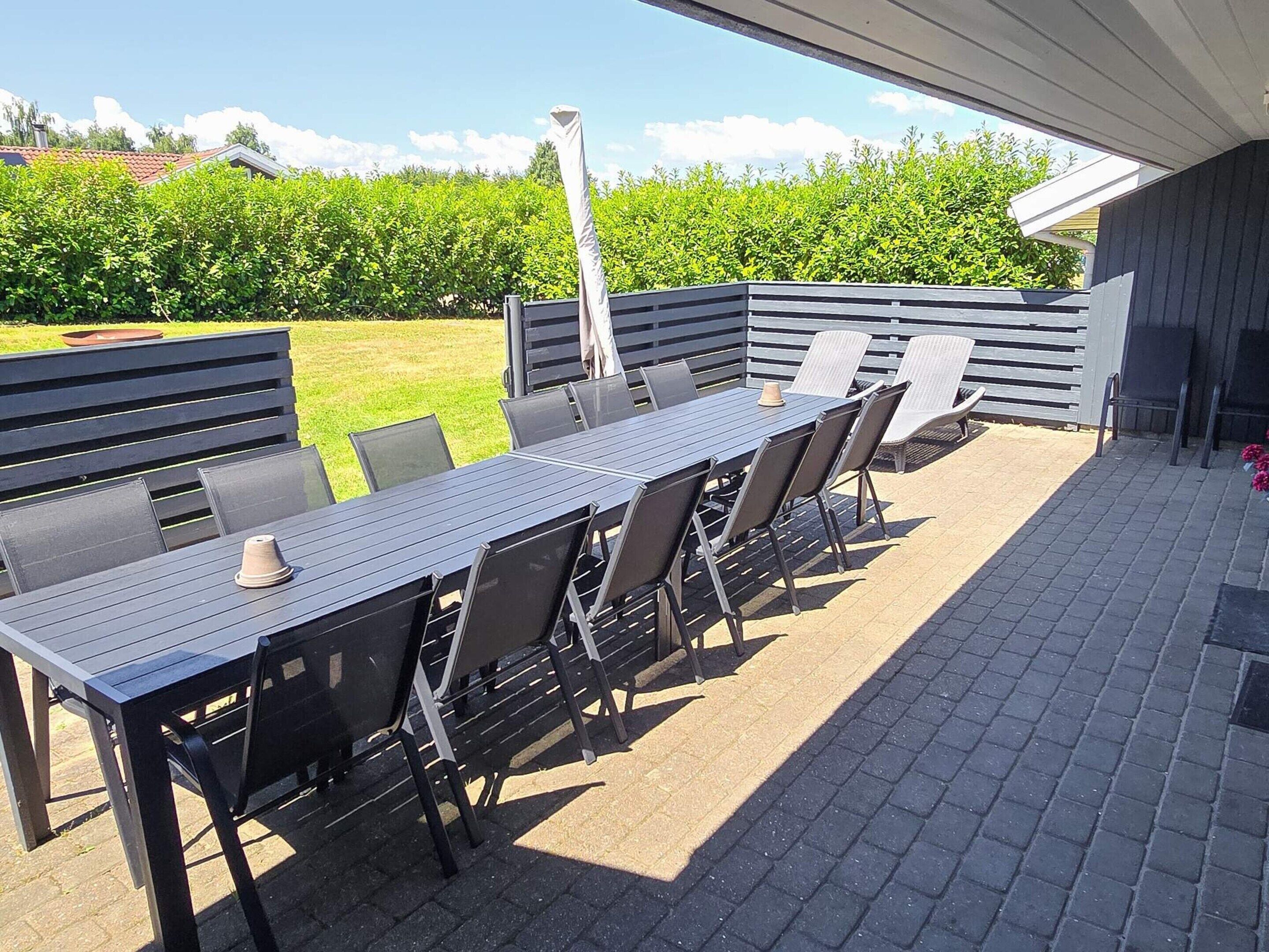 An outdoor patio with a long table and chairs, surrounded by a low fence and a hedge.