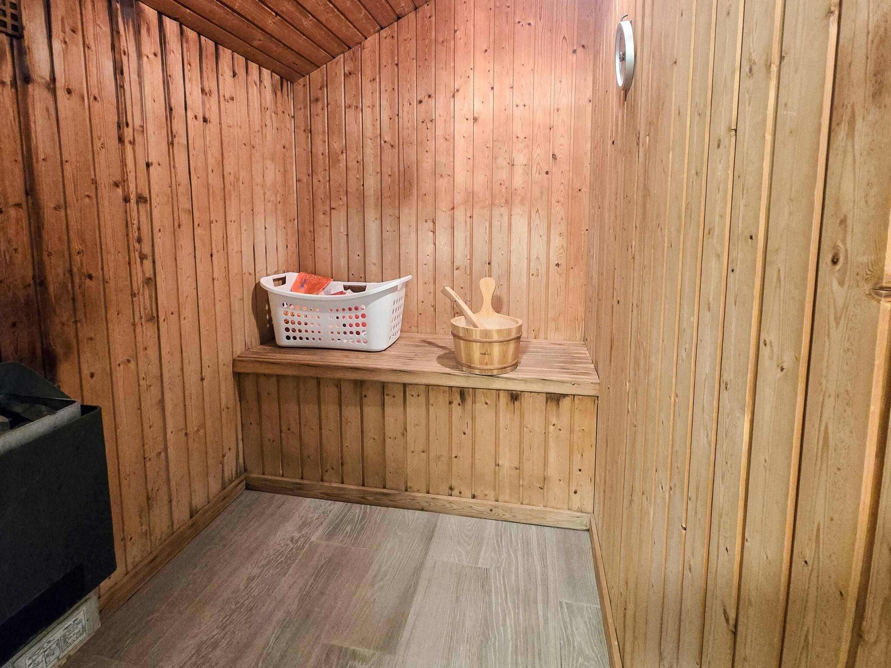 Et sauna-rum med trævægge, en bænk, en træspand og en hvid vasketøjskurv.