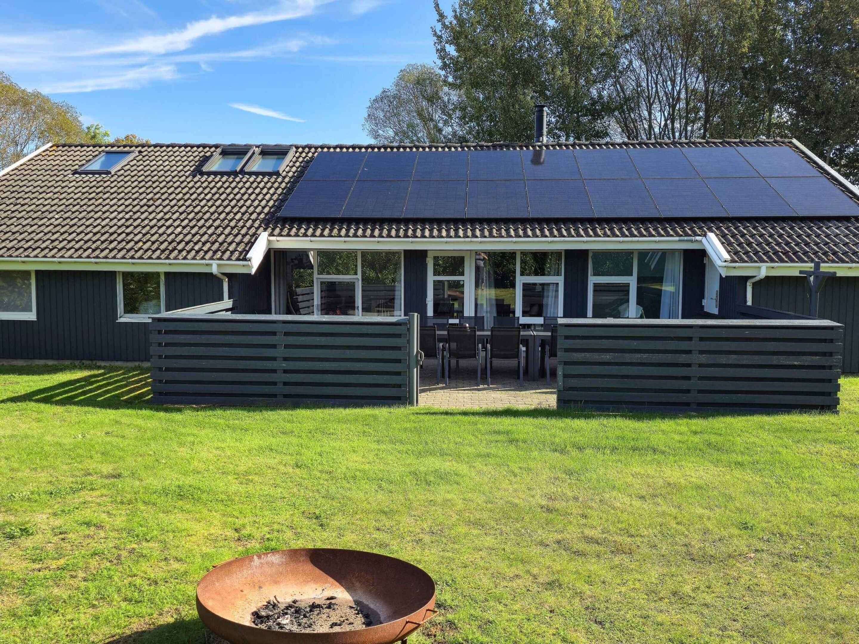 Une maison avec un toit  équipé de panneaux solaires, un patio couvert et un foyer extérieur dans le jardin.
