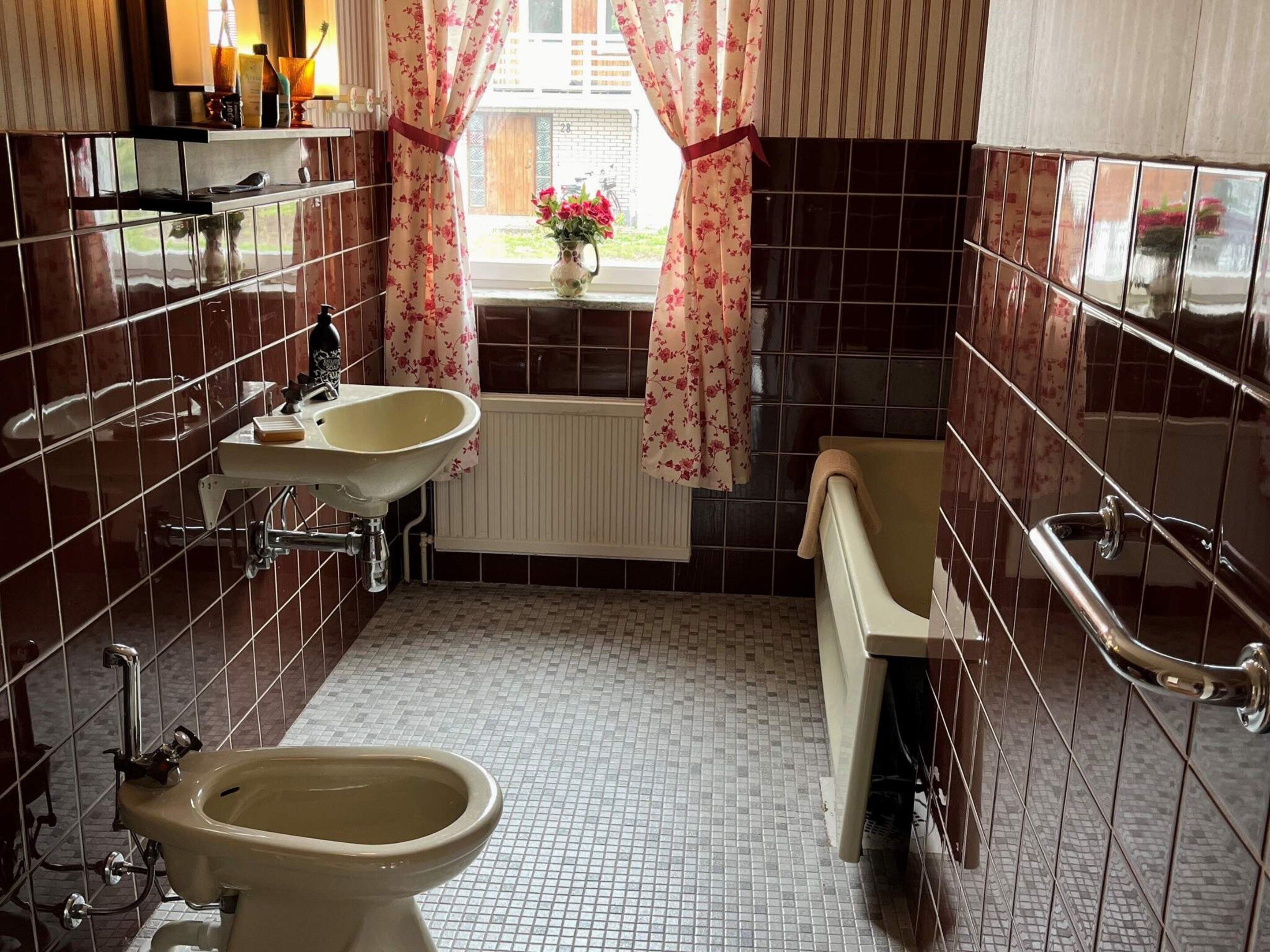 A bathroom with a bidet, a sink, a mirror, and a window with curtains.