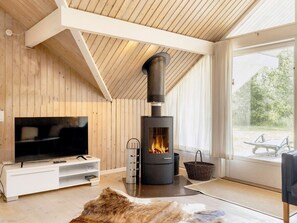 A modern living room with a large TV, a black wood-burning stove, and a view of the outdoors through a large window. - 6 Person Holiday Home in Rodby (Rodby)