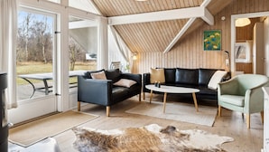 A spacious living area with a black leather sofa, a round coffee table, and a light green armchair. A wooden ceiling and large windows provide natural light and views of the outdoors.