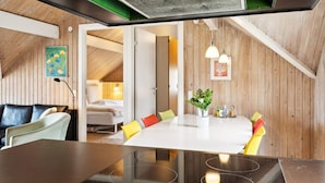 A modern kitchen with a central island, a dining area with colorful chairs, and a visible bed in the adjacent room.