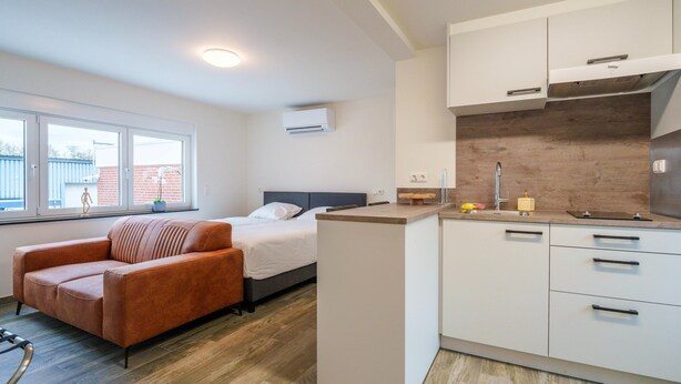 A modern hotel room with a bed, a sofa, a kitchenette, and a window with a view of a building. - Cosy Apartment Near Center (Landgraaf)