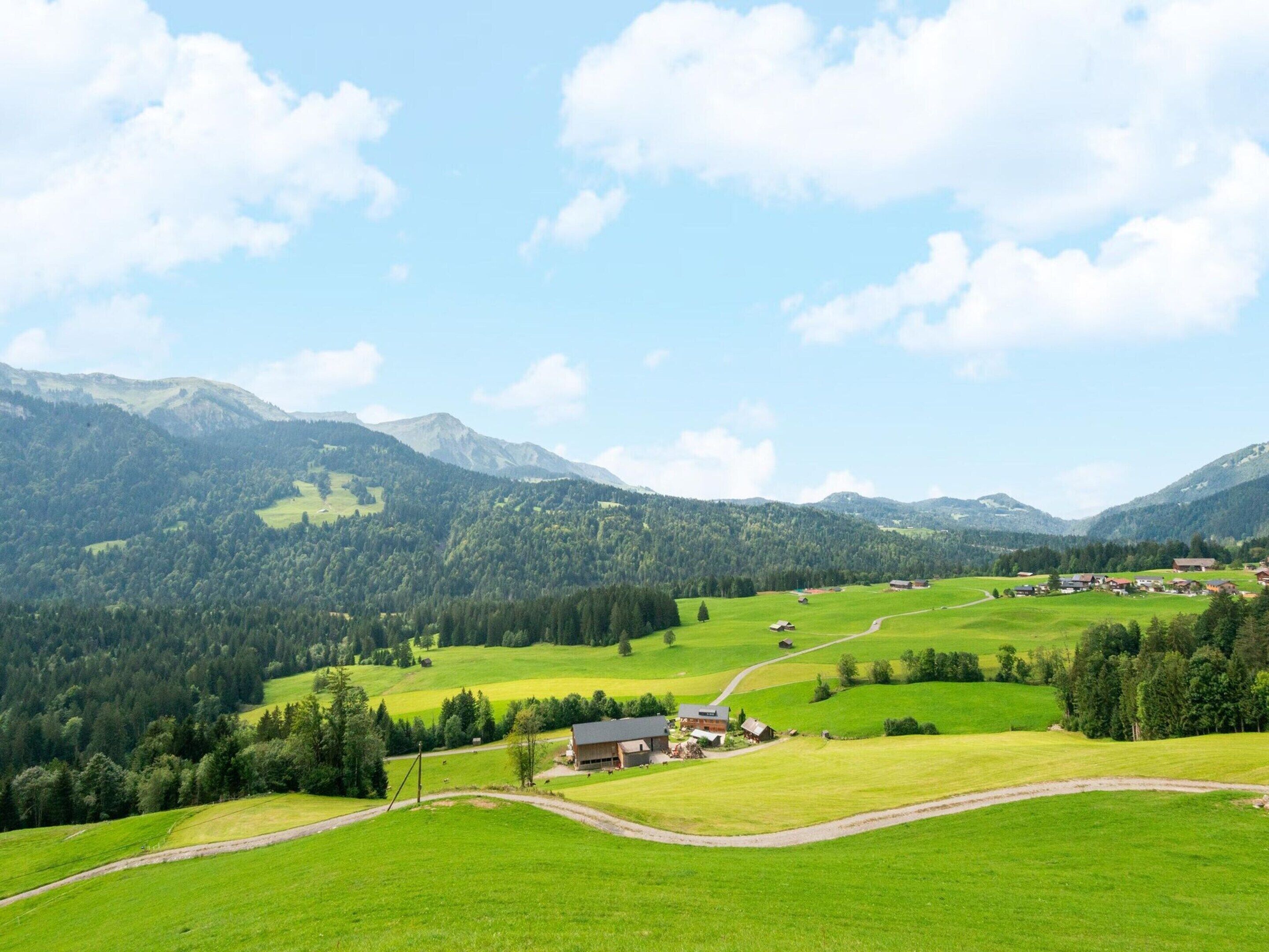 A rural landscape with rolling green fields, a winding road, and a small cluster of buildings in the valley.