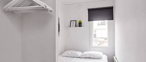Habitación pequeña con cama individual, pared blanca, ventana, radiador y repisa con una planta.