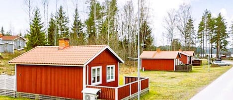 A red wooden cabin with a porch and a small attached shed, surrounded by trees and a road.