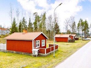 Una cabaña roja de madera con porche y un cobertizo pequeño adosado, rodeada de árboles y una carretera.