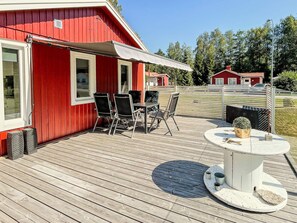 A red building with a covered patio area featuring a table and chairs, a small round table with a plant, and a fenced yard with another red building in the background. - 6 Person Holiday Home in Virserum (Virserum)