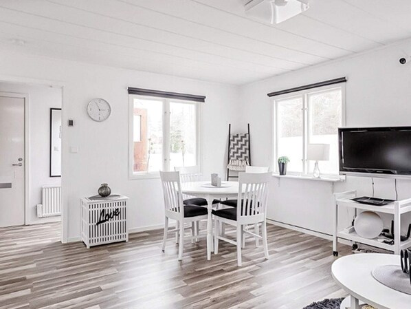 A modern, white-themed living room with a dining area, a flat-screen TV, and a fireplace.