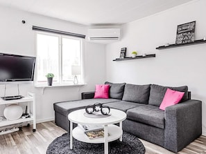 Un salon moderne avec un canapé d’angle gris, une table basse blanche, un téléviseur à écran plat et une fenêtre équipée de stores.