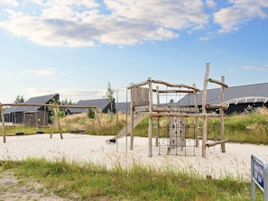 A playground with swings and a slide, surrounded by sand and grass, with buildings in the background. - 5 Star Holiday Home in Romo (Rømø)