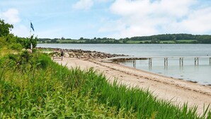 Ein Strand mit einem hölzernen Steg, der sich über ruhiges Wasser erstreckt, umgeben von Grünpflanzen und einem klaren Himmel.