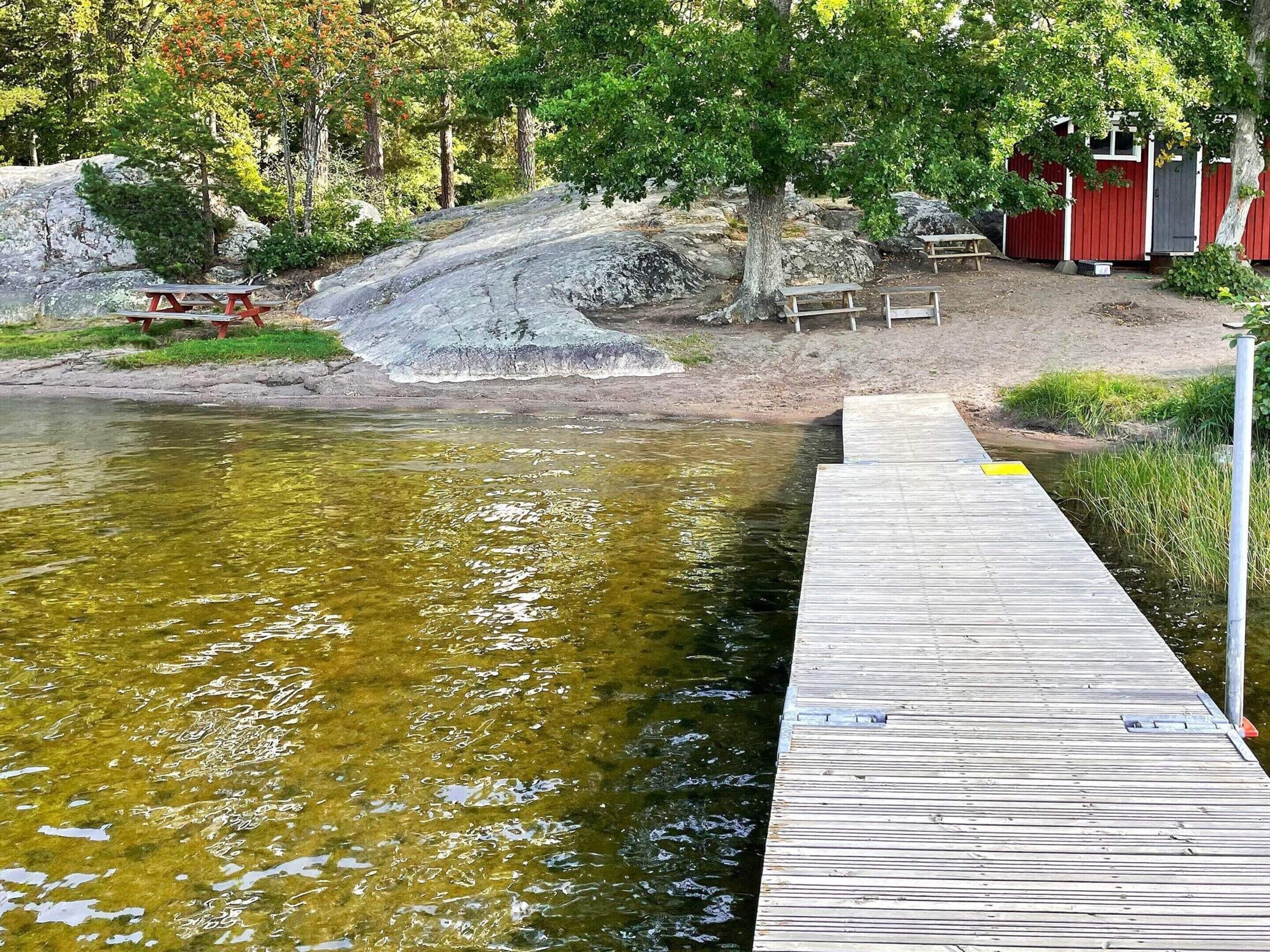 A wooden dock extending into a clear body of water, surrounded by trees and a red building with a gray door.