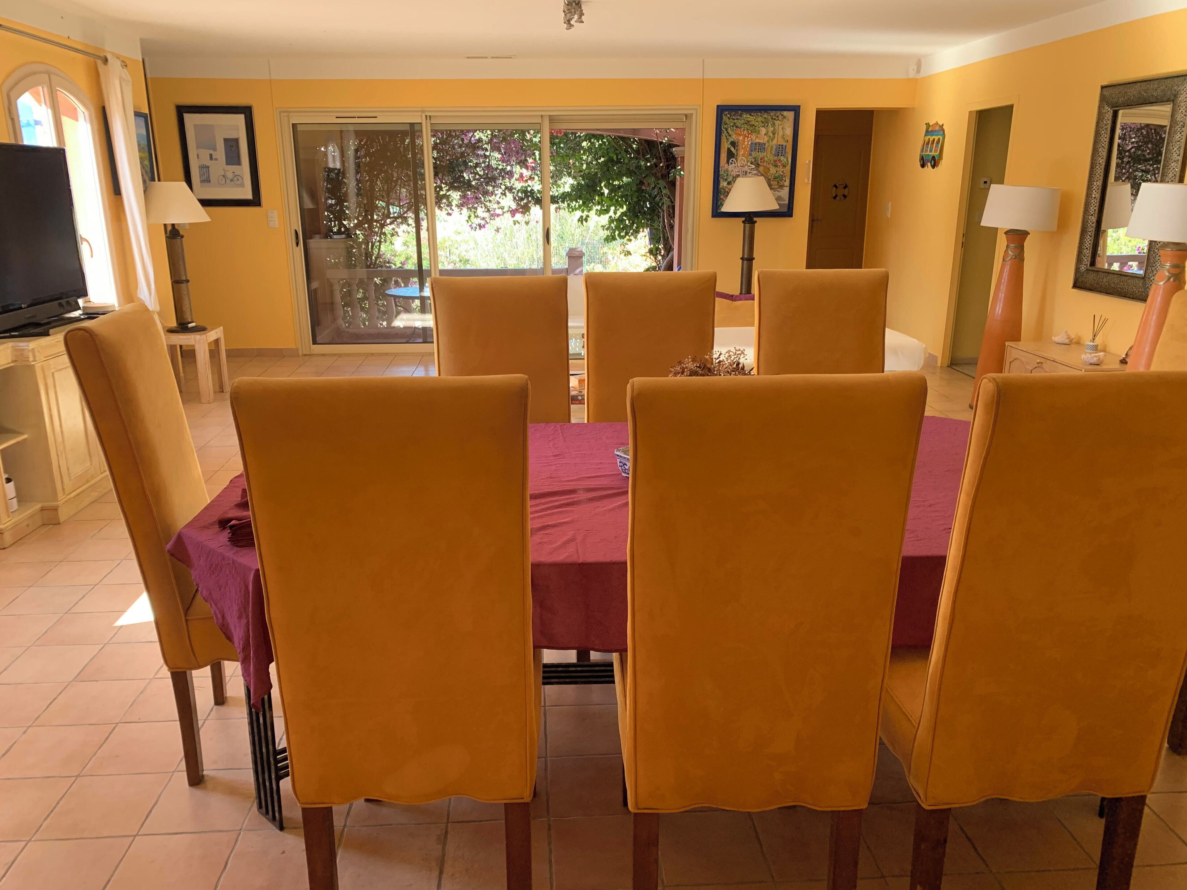 Un coin repas avec une table dressée pour six personnes, entourée de chaises, dans une pièce aux murs jaunes et dotée d’une grande fenêtre.
