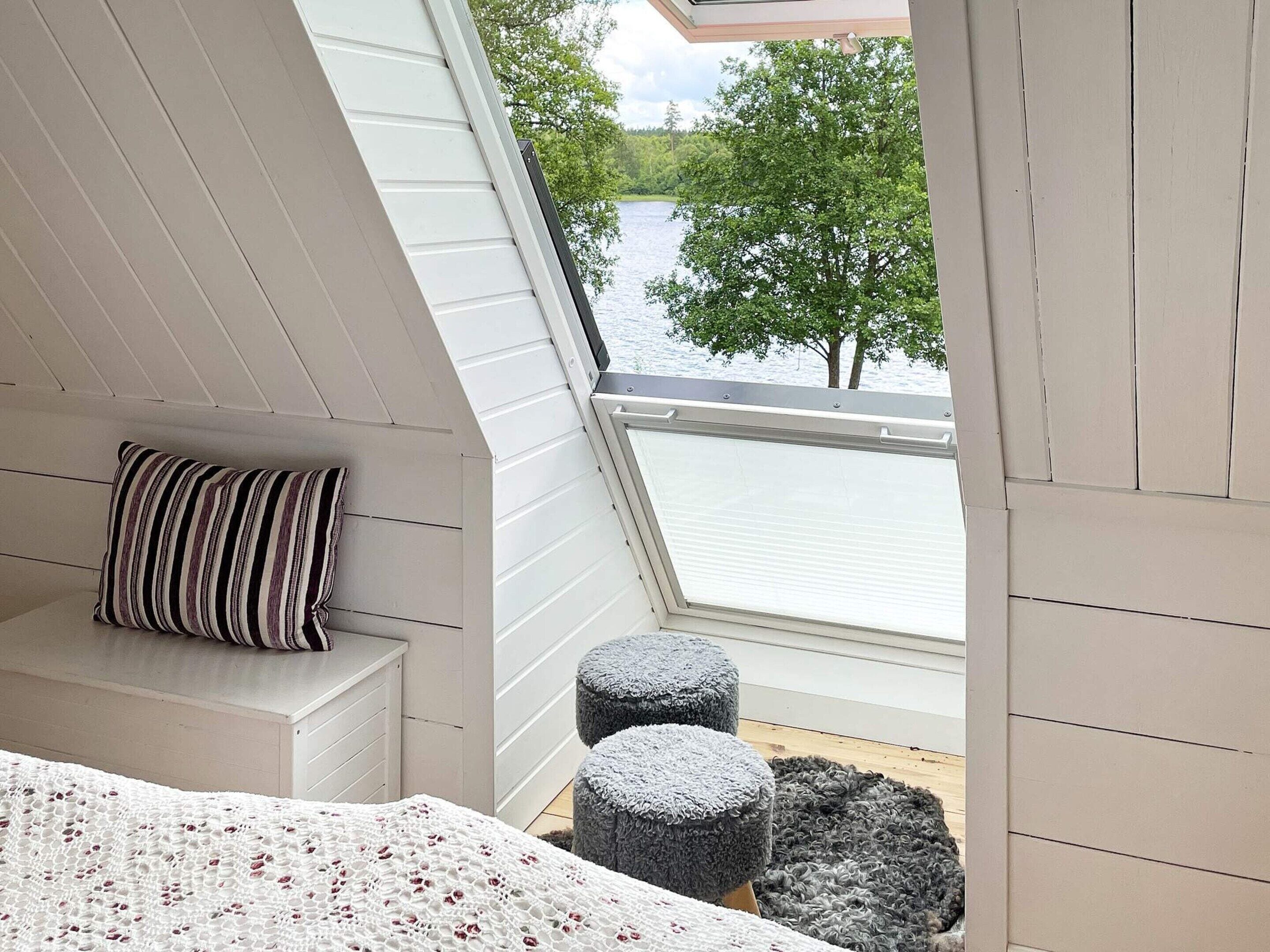 A bedroom with a sloped ceiling, a window overlooking a lake, a bed with a white blanket, a striped pillow, and two grey ottomans.