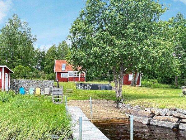 A lakeside cabin with a dock extending into the water, surrounded by lush greenery and a red building in the background. - 4 Star Holiday Home in Osby (Osby)