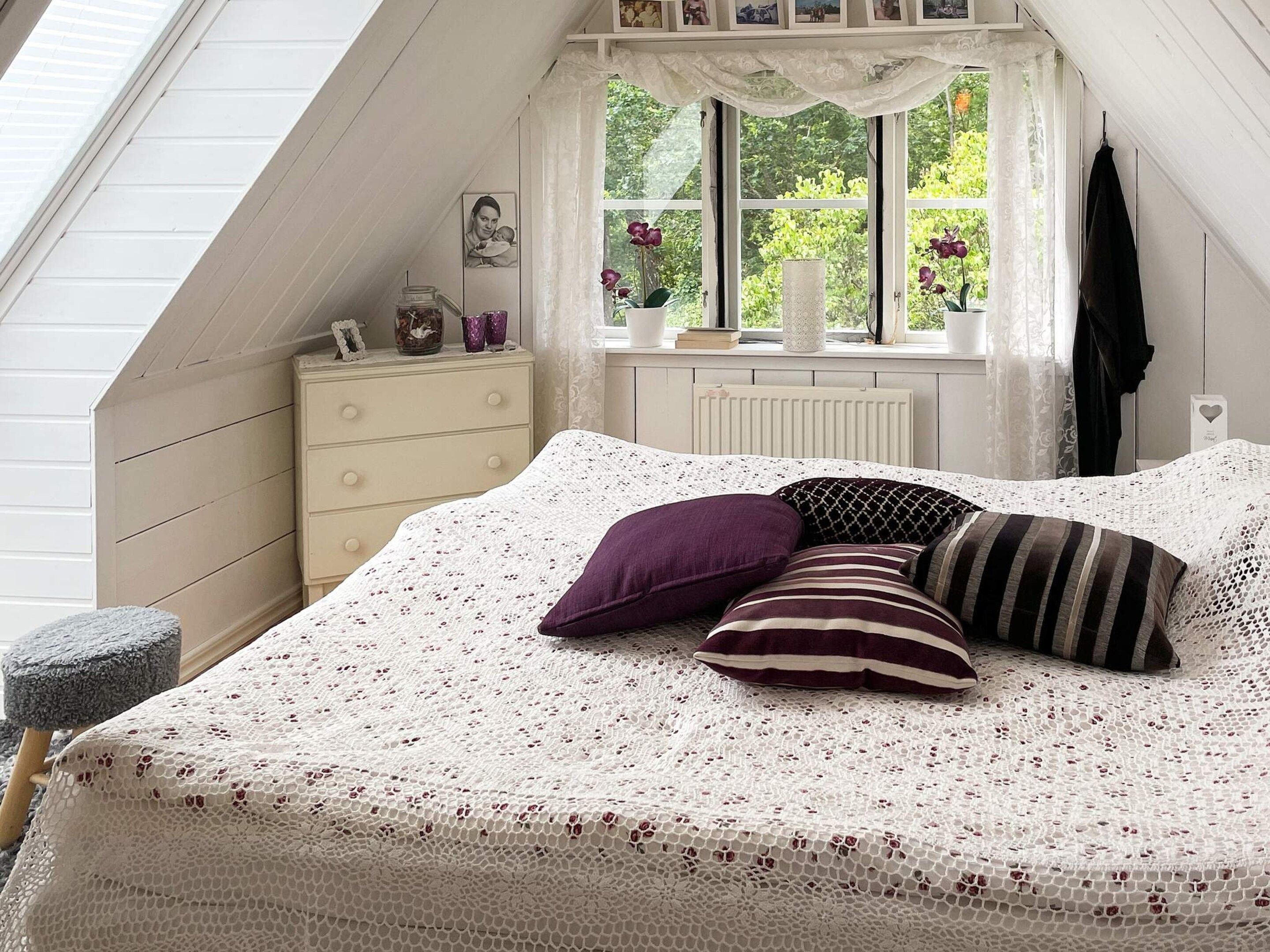 A bedroom with a sloped ceiling, a bed with a white knitted bedspread, a dresser, a window with lace curtains, and a radiator.