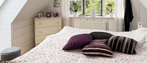 A bedroom with a sloped ceiling, a bed with a white knitted bedspread, a dresser, a window with lace curtains, and a radiator.