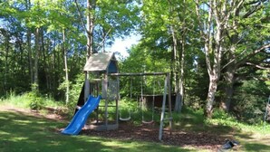 Un terrain de jeux avec un toboggan et des balançoires, entouré d’arbres.